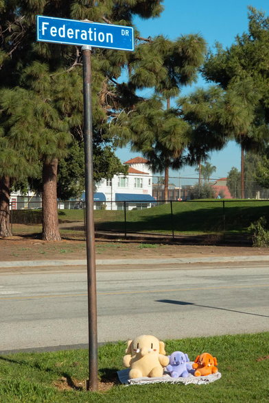 Photo of the Creature, Strawberry, and Blueberry Mastodon stuffed toys sitting outdoors by the sign for Federation Drive during a bright, sunny afternoon. From image-left to image-right, the Creature, Blueberry, and Strawberry are sitting on a folded beach towel that's been placed over some grass in the foreground. Next to them, to the right of the Creature, is a tall pole, atop which is the nameplate for Federation Drive. In the midground is Federation Drive, empty of any cars, and further in the background is a golf course, pine trees, and some distant buildings.