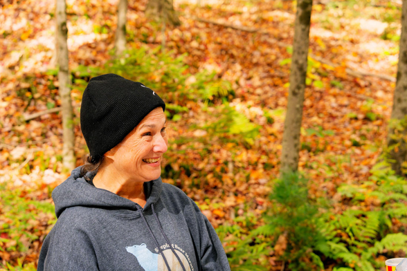 A smiling woman wearing a grey hoodie and a black hat looks right and behind her is a forest floor with a few ferns and many fallen leaves