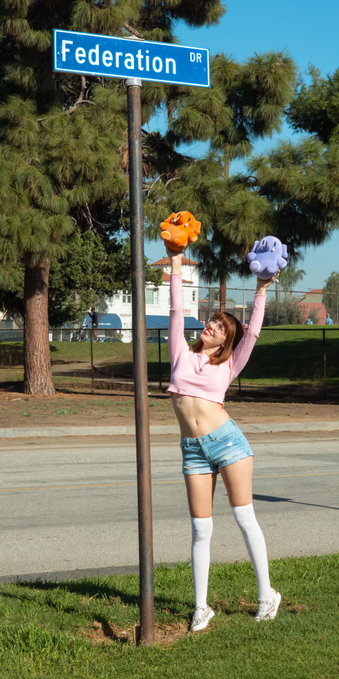 Photo of Lety with Strawberry II and Blueberry II, Mastodon stuffed toys, outdoors by the sign for Federation Drive during a bright, sunny afternoon. From top to bottom, Lety is shown wearing a pink long-sleeve crop top, light blue denim shorts, white thigh-high socks, and white-and-floral-print sneakers. She's tiptoeing on some grass in the foreground, standing next to a pole with her back arched to reach her arms in the air as high as possible. In Lety's right and left hands are Strawberry II and Blueberry II, respectively, raised as close as she can manage to the nameplate of Federation Drive on top of that pole. Further in the background is a golf course and some distant buildings, but right behind her is Federation Drive, devoid of cars, which is completely unrepresentative of everyone who slowed down to stare at her over the several minutes she spent posing for these photos.