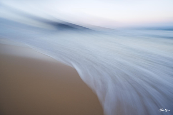A sweeping wave crashes across a beach with distant hills. overall bright but muted colours. abstract coastal scene