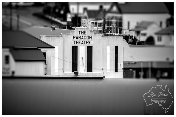 Black and white tilt shift photograph of the Art Deco facade of "The Paragon Theatre" in Queenstown, Tasmania. The name is prominently displayed above the vertical black and white stripe entrance.  The theatre is surrounded by older residential and commercial buildings, with a blurred foreground rooftop creating a shallow depth of field effect.