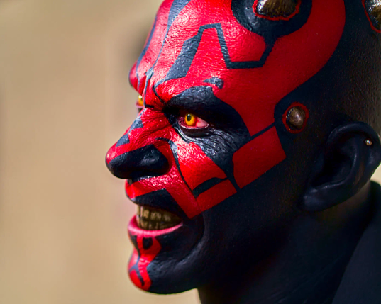 Profilaufnahme von Darth Maul, mit seinem charakteristischen Kopf in Rot und Schwarz und einem hassverzerrten Lächeln im Gesicht. Die Zähne sind grau und ungepflegt, die gelben Augen stechen aus dem Gesicht hervor. 