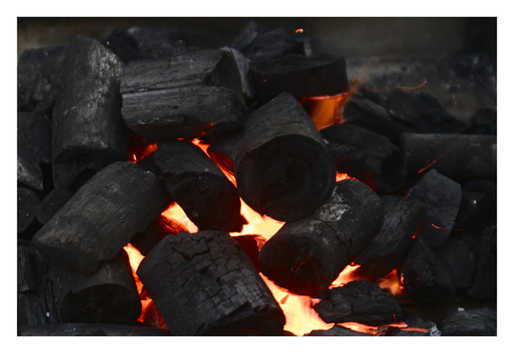 A pile of stacked, dark lump charcoal pieces actively burning. Intense orange and yellow embers radiate brilliant heat from the crevices and gaps between the black logs, with faint sparks visible in the air above the glowing core. - Google Gemini Latest