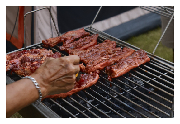 A hand brushing red marinade onto several pieces of raw, thick-cut meat laid across a metal barbecue grill outdoors. The grill sits over hot charcoal, and the background shows a blurred camping tent and grassy ground. - Google Gemini Latest