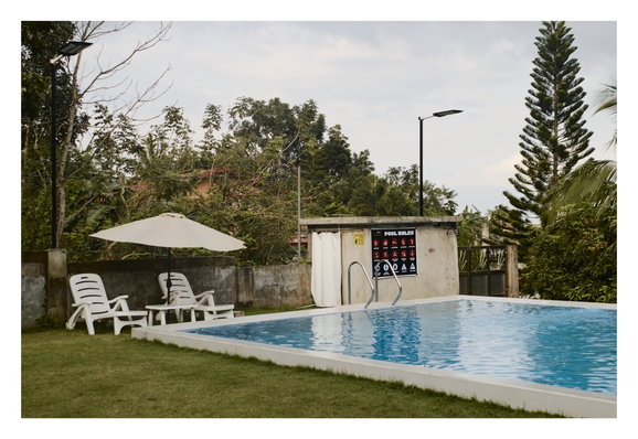 An outdoor swimming pool, featuring bright blue rippling water, is shown adjacent to a well-maintained lawn. Two white plastic loungers and a beige umbrella sit on the grass beside a concrete wall, which holds a "Pool Rules" sign. Lush, dense tropical foliage and an overcast sky dominate the background. - Google Gemini Latest