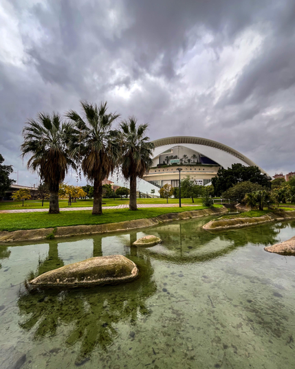 Un cielo nublado se cierne sobre una estructura arquitectónica moderna con un techo curvo (el Palau de les Arts), rodeada de exuberante vegetación y palmeras, reflejadas en un canal de agua.