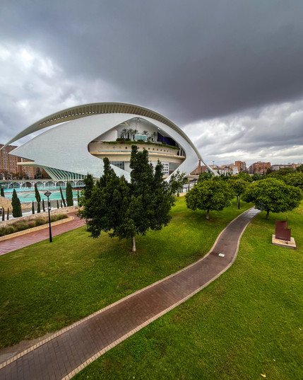 Edificio futurista con un diseño curvo (el Palau de les Arts) en medio de un exuberante césped verde y árboles bajo un espectacular cielo nublado, que ofrece un contraste moderno pero natural.