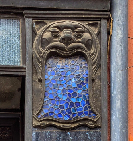 This image showcases an architectural detail of a building façade. At the centre is a decorative stone relief featuring the stylised face of a lion. The lion’s mouth is open, forming an arch around a small, rectangular stained-glass window. The stained glass is composed of irregular, mosaic-like pieces in shades of blue, creating a striking geometric pattern. At first glance, it looks as if the lion is puking water. The surrounding area of the building appears to be made of dark stone and there‘s a rainwater pipe visible next to the lion.