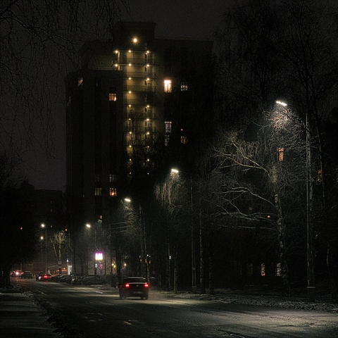 Late evening. In the foreground, a road stretches into the distance, its surface dotted with the moving lights of cars. Glowing streetlamps line the roadside, their light illuminating the swirling snow and the delicate branches of nearby trees. In the background, a modern multi-story building rises, its windows and balconies aglow with warm light.