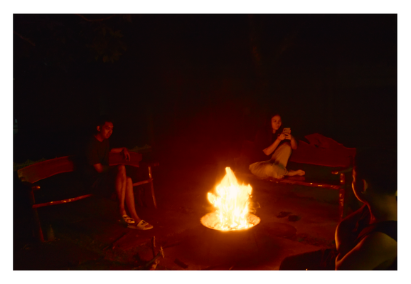 Friends gathering around a bright campfire. In the center, flames rise from a fire pit, casting a warm orange glow that illuminates the surroundings against the pitch-black darkness. - Google Gemini 3 Pro Preview