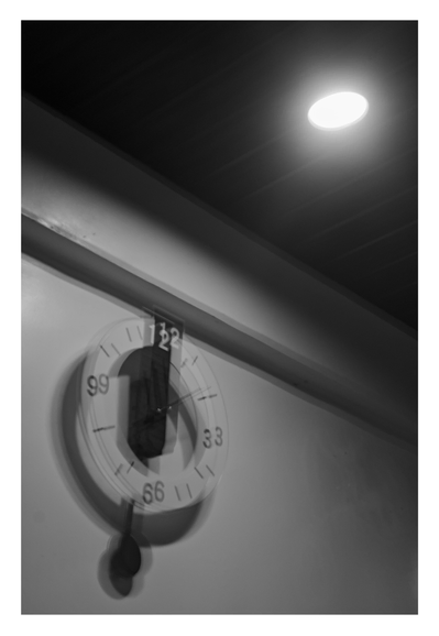 A wall clock and a ceiling light. The image has a distinct motion blur or "shaky" effect, causing the numbers on the clock face to appear doubled (resembling 99, 33, and 66) and giving the object a vibrating appearance. Above the clock, a bright, circular recessed light shines intensely against the dark ceiling - Google Gemini Latest