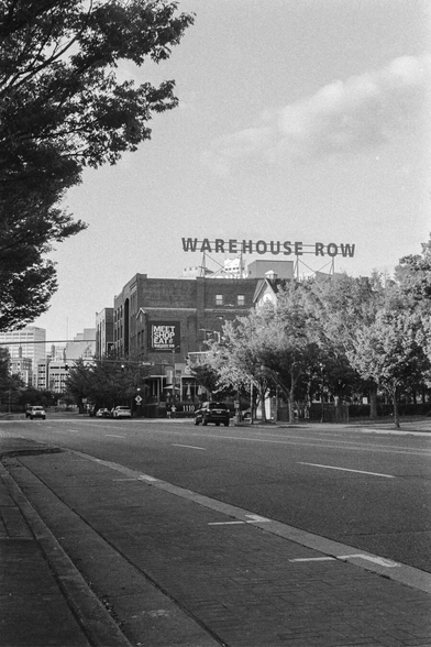 Black-and-white photograph of a wide city street with low brick warehouse buildings. A large rooftop sign reading “WAREHOUSE ROW” rises above the buildings in the distance. The street is mostly empty, with a few cars, sidewalks, trees lining the road, and a quiet urban atmosphere.