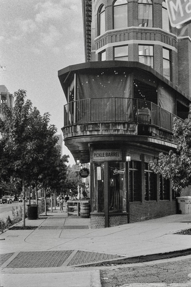 Black-and-white photograph of a corner brick building with rounded architecture and large windows. A sign reading “Pickle Barrel” is visible at street level. Trees line the sidewalk, a few people are seated outside in the distance, and the scene feels calm and understated in an urban neighborhood