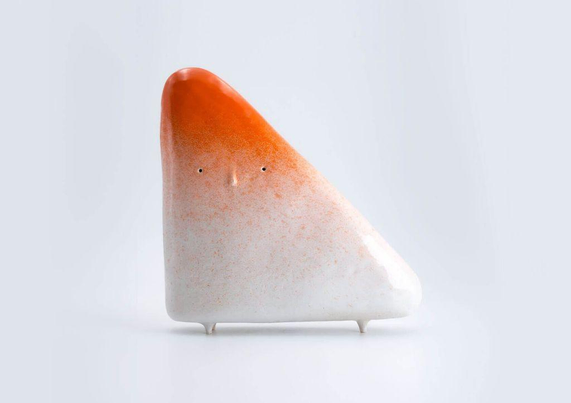 A whimsical ceramic sculpture resembling a face, with a glossy orange top and matte white bottom, resting on three small feet.