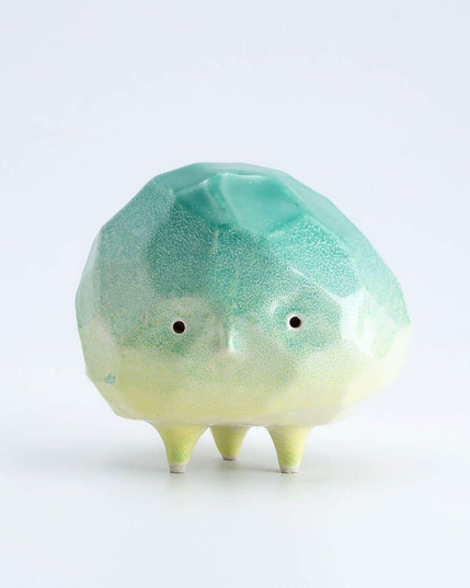 A whimsical ceramic creature with a turquoise and light green gradient, featuring two small eyes and three short legs. 
