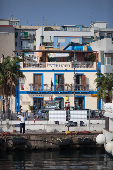 A brightly painted building with the sign “PETIT HOTEL” is visible, featuring balconies with plants and flags. In the foreground, a person stands on a dock by the water, with boats and palm trees nearby.
