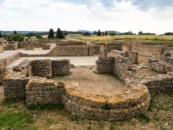 Color photo of the low ruin walls of Roman houses with some of the mosaic floors still intact, set in a Catalonian landscape with rolling fields and hills in the background. 