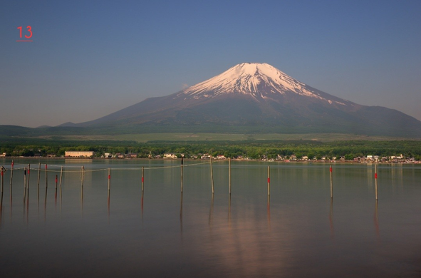 Photo couleur du mont Fuji depuis Yamanaka. En arrière-plan, le mont Fuji avec le sommet enneigé. Au premier un lac où se reflète vaguement la montagne. De l'autre côté du lac, on distingue les bâtiments d'une petite ville longeant ce lac. Le ciel est bleu et il n'y a pas un nuage.