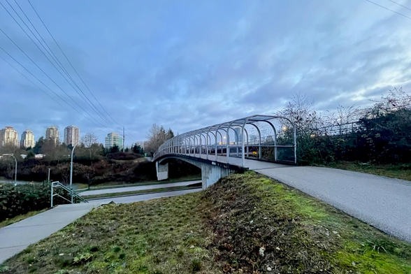 The image shows a pedestrian bridge with a covered walkway under a cloudy sky. In the background, there are high-rise buildings and power lines.