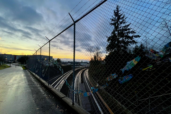 The image shows a chain-link fence with barbed wire at the top, over a set of train tracks. The sky is filled with colourful clouds at sunset.