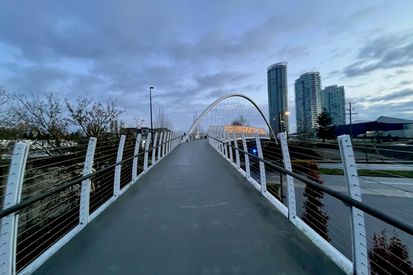 The image shows a pedestrian bridge with a curved arch in the middle of the span. The bridge is in an urban setting with high-rise buildings in the background.