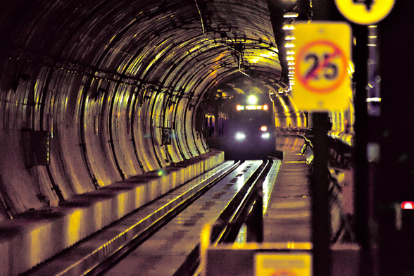 highly stylized photo of an underground train in a circular tunnel. It approaches toward the viewer and casts the tunnel in yellow light with purple shadows
