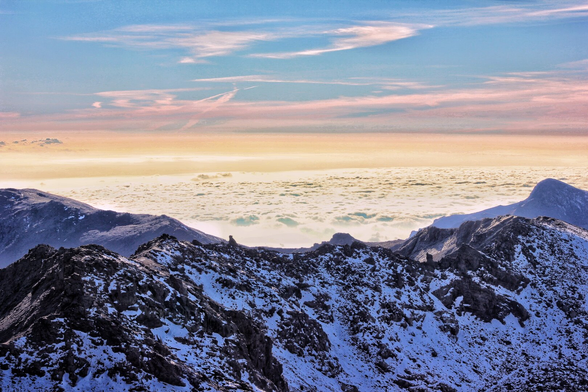 A breathtaking view from a high mountain peak looking down over a thick blanket of white clouds. The foreground features rugged, dark rocks dusted with snow. The horizon glows with the warm, pastel colours of a setting or rising sun, transitioning from golden yellow to soft pink and blue in the sky.