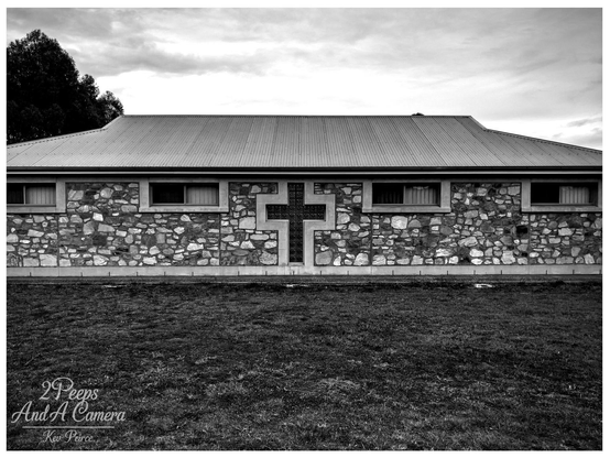 A stark, symmetrical black and white photograph of the side hall or auxiliary building of Manaranga Church.

The structure features a low pitched roof of corrugated iron and a rugged exterior wall constructed from varied, irregular fieldstones.

Centred on the wall is a large, recessed cross made of glass block or heavily framed stained glass, flanked by two horizontal windows on either side. A grassy foreground sits beneath a cloudy sky.