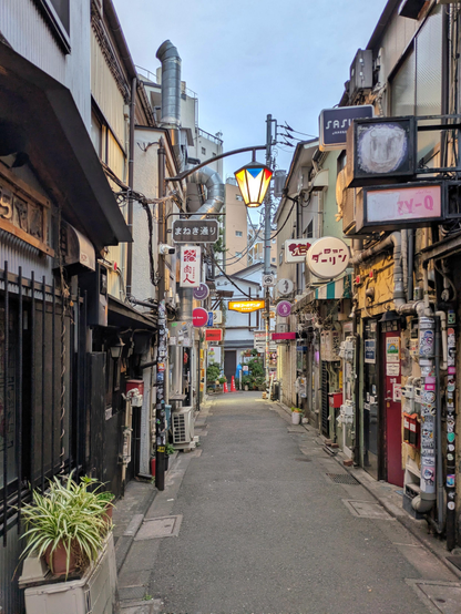 A narrow Japanese alley lined with small bars and restaurants, viewed straight down the street. Old buildings crowd both sides, covered with pipes, vents, stickers, and hanging signs in Japanese. A decorative streetlamp hangs overhead, and the alley is empty, creating a quiet, early evening atmosphere.