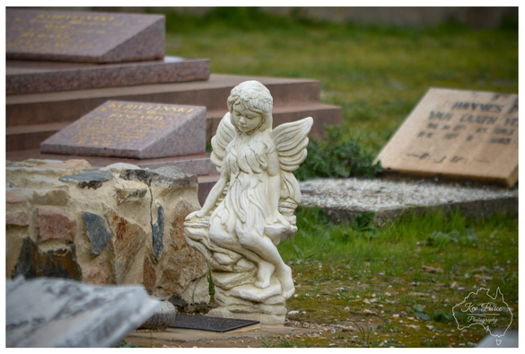 A colour photograph of a small, white stone statue of a seated angel with wings, resting on a rocky base in a cemetery.  The angel is carved in a delicate style, looking down. In the middle ground to the left, there is a large, rough hewn stone border, and behind the angel, several modern granite grave markers with sloped faces are visible, some inscribed with gold lettering. The ground is covered in short grass.