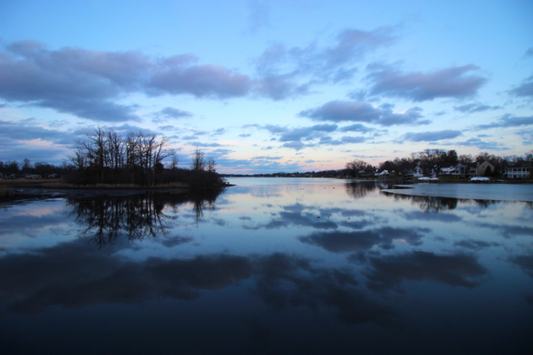 Photograph of a lake at twilight with clouds above reflected in the mirror-smooth water below. There is a small island to the left and some ice on the surface to the right.