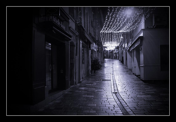 A narrow cobblestone street draped in night settles into a hushed, almost cinematic stillness. The pavement is slick — the stones catching and scattering the soft glow of a canopy of tiny lights strung overhead like a low, luminous sky. Old shopfronts and shuttered windows line both sides, their facades sinking into gently deep shadows; the architecture leans inward, guiding the eye down the diminishing perspective to a faintly lit vanishing point. A thin pair of tram or drainage rails runs down the center of the road, their dark lines becoming a rhythmic spine through the scene.
Rendered in monochrome, the image emphasizes texture and contrast: the glossy reflections on the wet stones, the grain of weathered walls, and the delicate sparkle of bulbs that halo with a mild, diffused radiance. There’s an intimate, slightly melancholic mood — as if the street has just closed for the night and is holding its breath, waiting for the next footstep. 