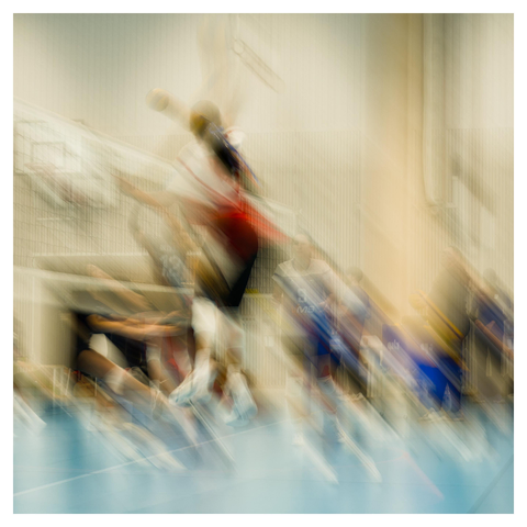 En fyrkantig bild på tjejer som spelar volleyboll. Bilden är tagen med lång slutartid och rörelse av kameran så allt är svepande suddigt. En tjej i röd tröja hoppar högt och slår nära nätet. Lagkamrater i blått tittar på och motståndare i mörka tröjor försvarar.