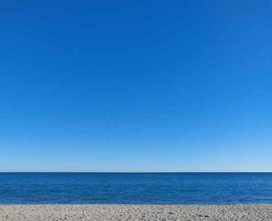 Photo d'un bord de plage déserte. Le sable fait une bande claire, il est recouvert de traces de pas. L'eau immobile est bleu marine et le ciel au dessus de la ligne d’horizon fait un dégradé du blanc jusqu’au bleu foncé.
