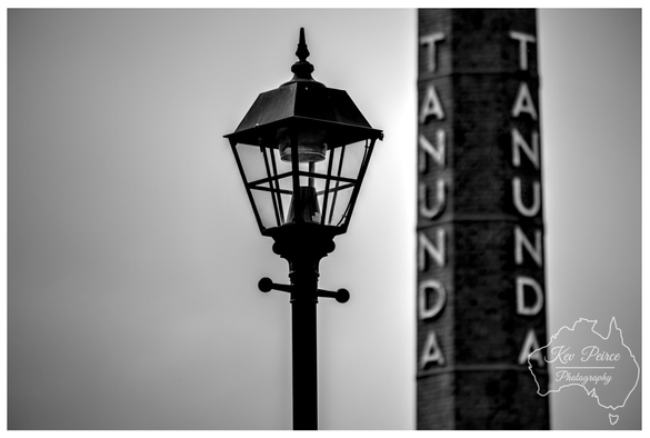 A monochromatic close up, low angle photograph featuring a classic, ornate, hexagonal street lamp (or lantern) silhouetted against a bright sky.  In the blurred background, out of focus, stands a tall hexagonal brick column or tower with the word "TANUNDA" written vertically down the sides. The focus is sharp on the details of the lantern.