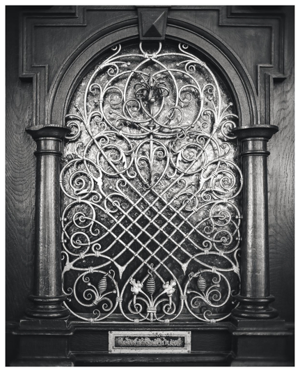 Das Bild zeigt ein schwarz-weißes, frontal aufgenommenes Detail einer Tür, genauer das verglaste Oberfeld mit einem schmiedeeisernen Ziergitter, eingefasst in einen massiven Holzrahmen.