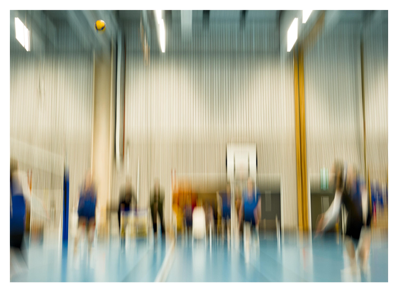 En liggande bild på ett volleyboll-lag som värmer upp. Allt är suddigt på grund av att kameran rört sig. Det enda som är i fokus är bollen högt uppe till vänster nära taket.