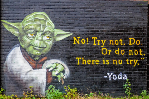 Eine Ziegelsteinwand mit einem Graffiti. Auf einer tief schwarzen Fläche ist die lächelnde Figur von Yoda aus Starwars aufgesprüht, wie er sich auf seinen Stock stützt. Rechts neben der Figur steht geschrieben: 'No! Try not. Do. Or do not. There is no try.' - Yoda