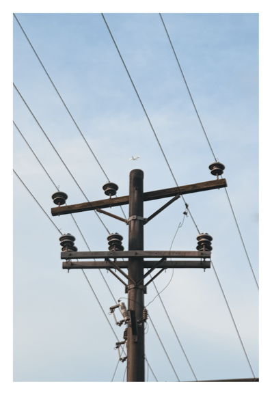 A wooden utility pole against a clear, pale blue sky. The pole features two horizontal crossarms holding several dark ceramic insulators, with multiple power lines stretching diagonally across the frame. High above the pole, a tiny, distant airplane can be seen flying through the sky. - Google Gemini 3 Pro Preview
