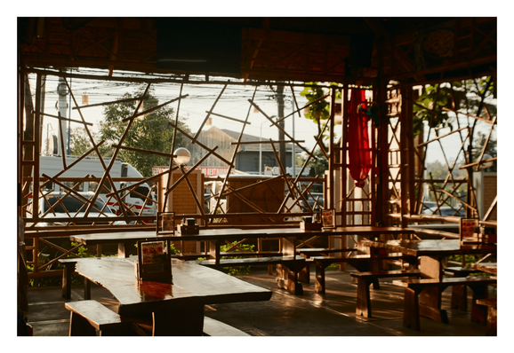 A rustic, open-air restaurant bathed in warm, golden sunlight. The structure is defined by a distinctive framework of crisscrossed bamboo poles that act as screens, offering a view of the street, trees, and vehicles outside.

Inside, sturdy, polished wooden tables and benches are arranged in rows. The low angle of the sun casts long shadows and bright highlights across the furniture and floor, creating a peaceful and inviting atmosphere. - Google Gemini 3 Pro Preview