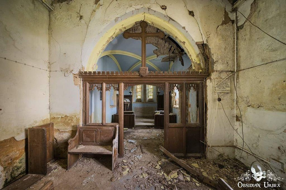 Image shows the decaying interior of an abandoned church. Ornate wooden decor leads to an altar space in front of stained glass windows.