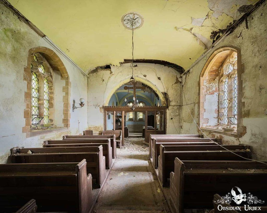Inside an abandoned church, peeling paint and rows of wooden pews create a haunting atmosphere. Light streams through arched stained-glass windows.