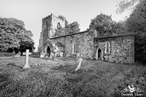 Black and white image of an abandoned church with a graveyard. Tower and arched doors and windows visible