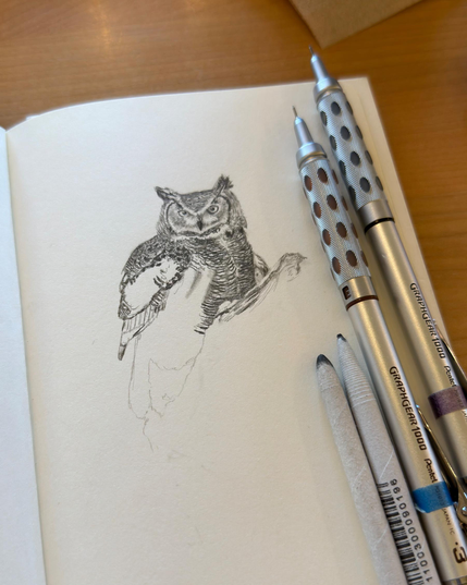 Open sketchbook on a wooden table with a partially finished graphite drawing of a great horned owl; two mechanical pencils and blending stumps lie beside the page.