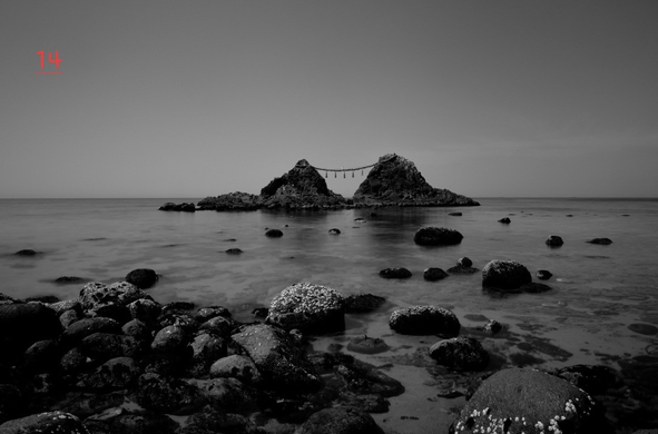 Photo noir et blanc d'un paysage marin. Au premier des rocher de diverses tailles, d'abord découverts puis peu à peu recouverts par la mer. Plus loin deux gros rochers reliés par une corde en paille de riz.