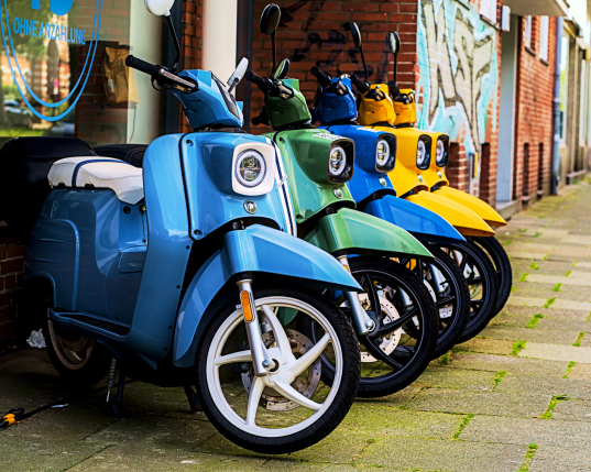 Vor einem Fenster stehen fünf bunte Roller in blau, grün und gelb am Rand eines Gehwegs. Auf dem Fenster steht in großen Buchstaben "Ohne Anzahlung". 
Es sind Elektroroller von Schwalbe, die hier verkauft werden. 
. 
Five colourful scooters in blue, green and yellow are parked in front of a window at the edge of a pavement. The window has ‘No deposit required’ written on it in large letters.
These are Schwalbe electric scooters that are sold here.
. 

