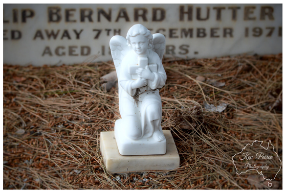 A small, white marble statuette of a kneeling angel is placed on a simple square base, resting on a bed of dry, reddish brown pine needles and earth.  The angel holds a cross to its chest. In the background, slightly blurred, is a white marble headstone with engraved text.