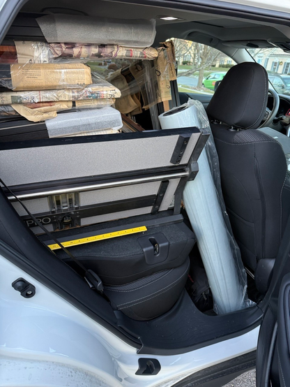 The view from a back passenger door of an SUV filled with frames, a mat cutter, and a large roll of paper.