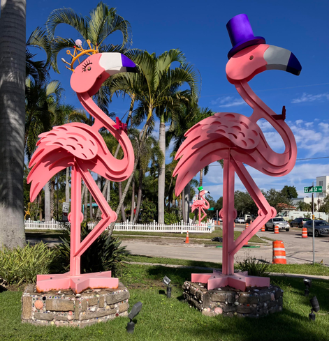 Two large flamingo statues, probably made of metal, and much taller than a person, stand on a lawn outside the Marietta Museum of Art & Whimsy (museum not in the photo). One flamingo wears a purple top hat, and the other wears a crown. You can see palm trees in the distance. 
