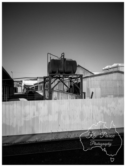 A striking black and white photo of an industrial scene. In the foreground, a low corrugated iron fence runs horizontally across the frame. Behind it, on a black metal frame, sit two large, rusty, cylindrical tanks.  Beyond the tanks are various corrugated iron sheds and industrial buildings, all under a vast, bright sky. A single railway track runs along the bottom of the image, below the fence line.